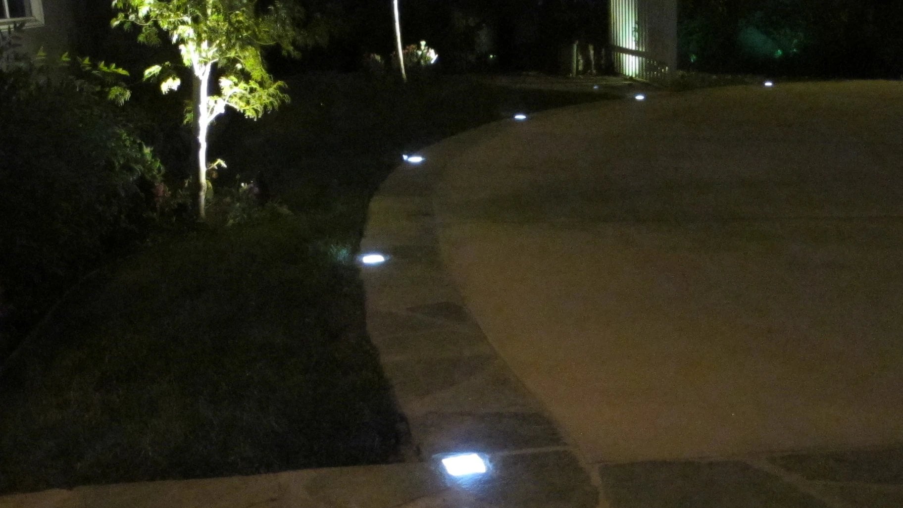 Gebogene Auffahrt zwischen Natursteinkante und Grünfläche bei Nacht, Lightstone LED-Pflastersteine säumen die Kante