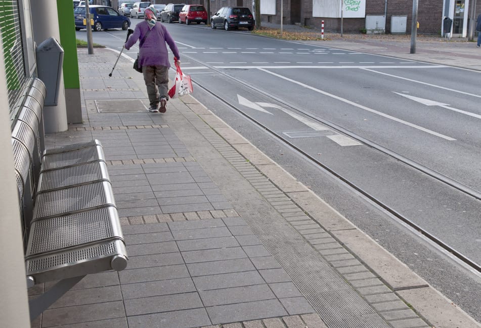 Haltestelle bei Tag mit weißem Leitstreifen in hellgrauem Pflaster, eine Frau mit Rollator