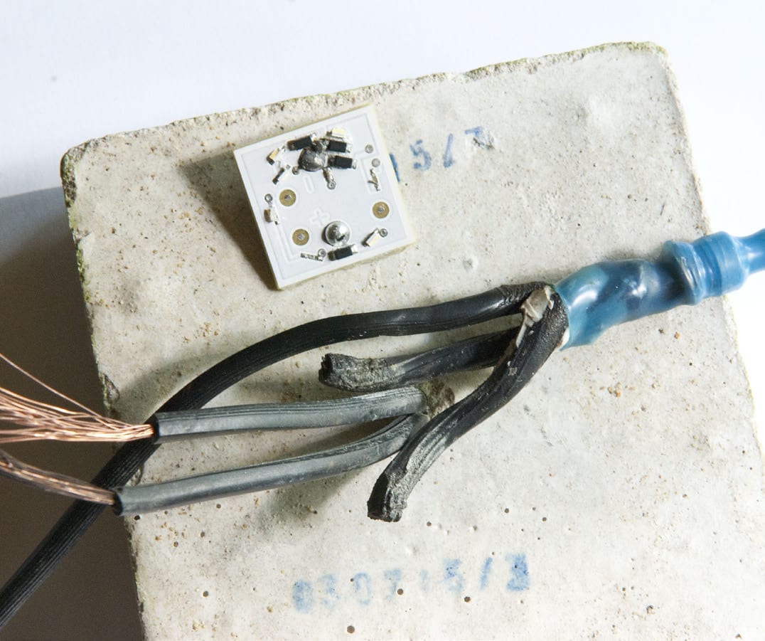 Geöffnete Leuchte auf einem Pflasterstein mit Platine, korrodierten Kupferlitzen und blauer Lötmuffe
