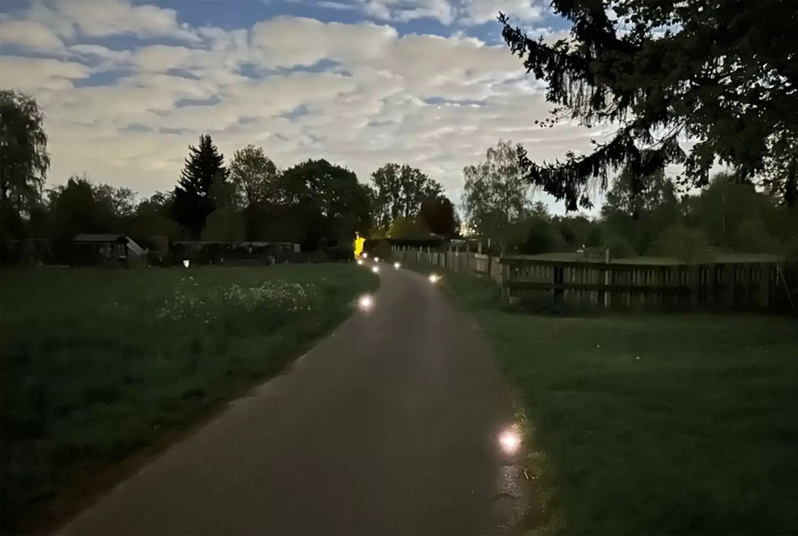 Radweg am Abend zwischen Wiese und Zaun, Road Studs leuchten am Wegrand